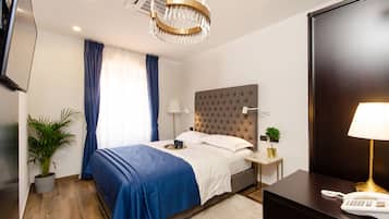 Habitación de hotel ordenada con cama, mesitas de noche, un escritorio con lámpara, televisor y planta en maceta.
