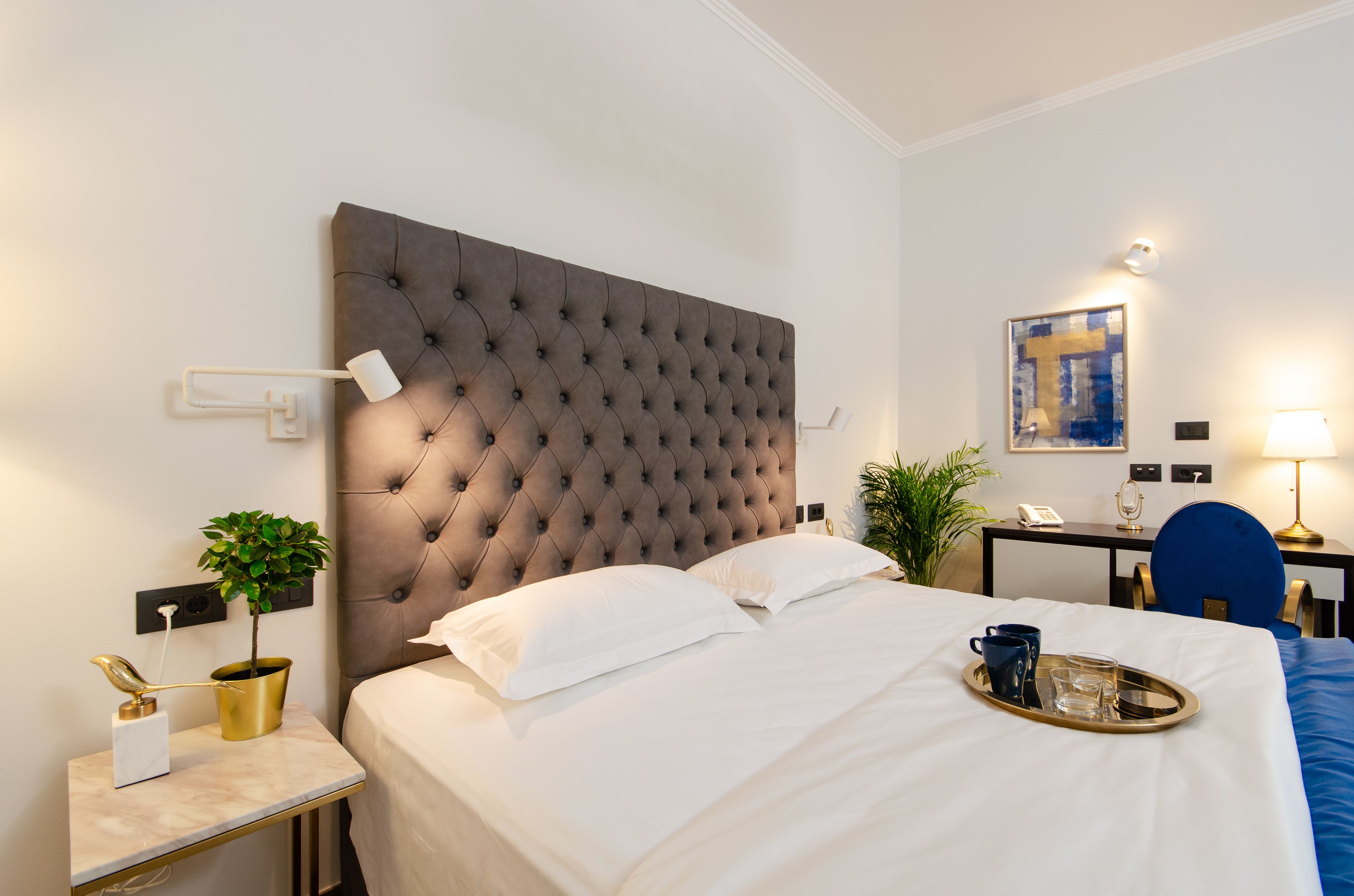 A hotel room with a tufted headboard, a bedside table with a plant, a tray with a mug and glasses, and a desk with a lamp.