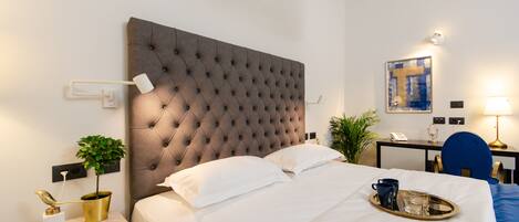 A hotel room with a tufted headboard, a bedside table with a plant, a tray with a mug and glasses, and a desk with a lamp.