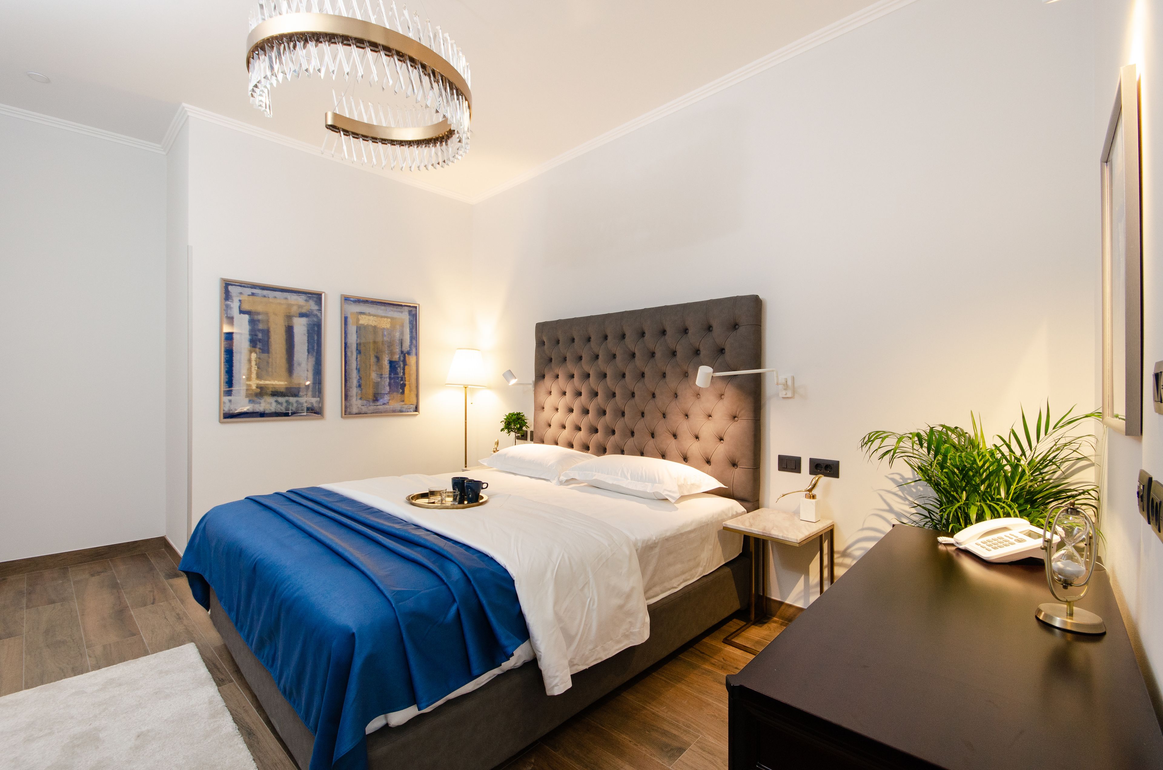 A modern bedroom with a tufted headboard, a bedside table with a lamp, a tray with a cup, and two framed artworks on the wall.