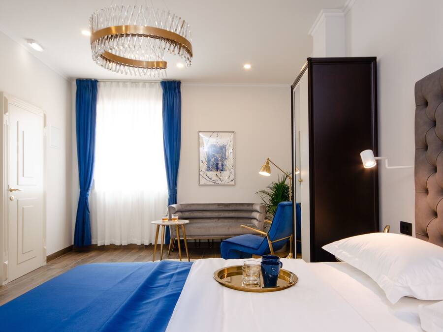A modern hotel room with a large bed, a blue accent wall, a chandelier, a sofa, and a dining table with a tray.