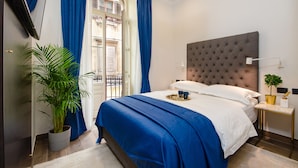 Un dormitorio ordenado con una cama grande, una planta en maceta, una mesita de noche con lámpara y vistas al exterior a través de una ventana con cortinas azules.