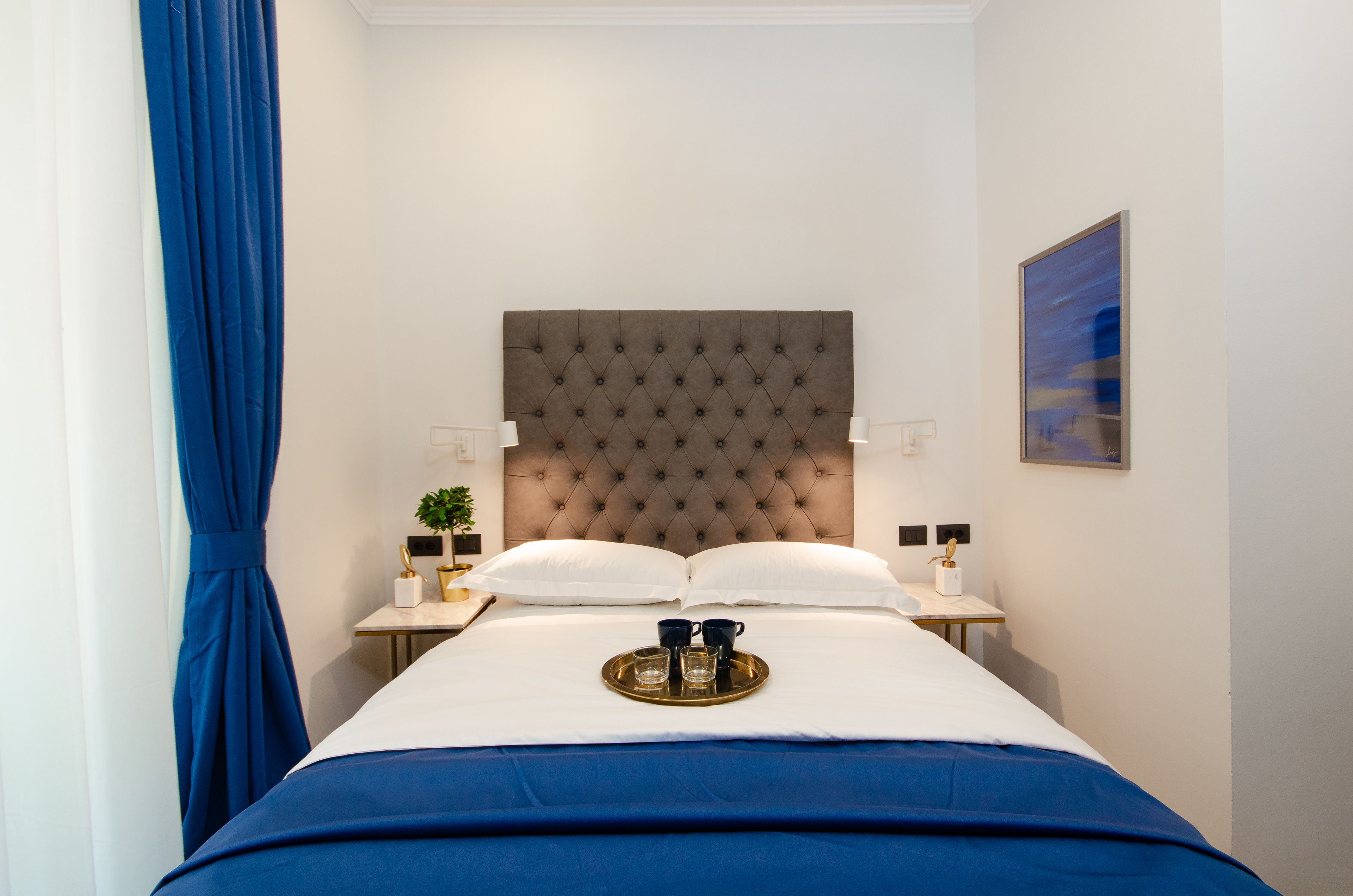 Un lit bien fait, doté d’une tête de lit capitonnée, deux tables de chevet avec des lampes, un plateau avec des tasses et un verre, et un tableau accroché au mur.