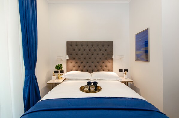 Un lit bien fait, doté d’une tête de lit capitonnée, deux tables de chevet avec des lampes, un plateau avec des tasses et un verre, et un tableau accroché au mur.