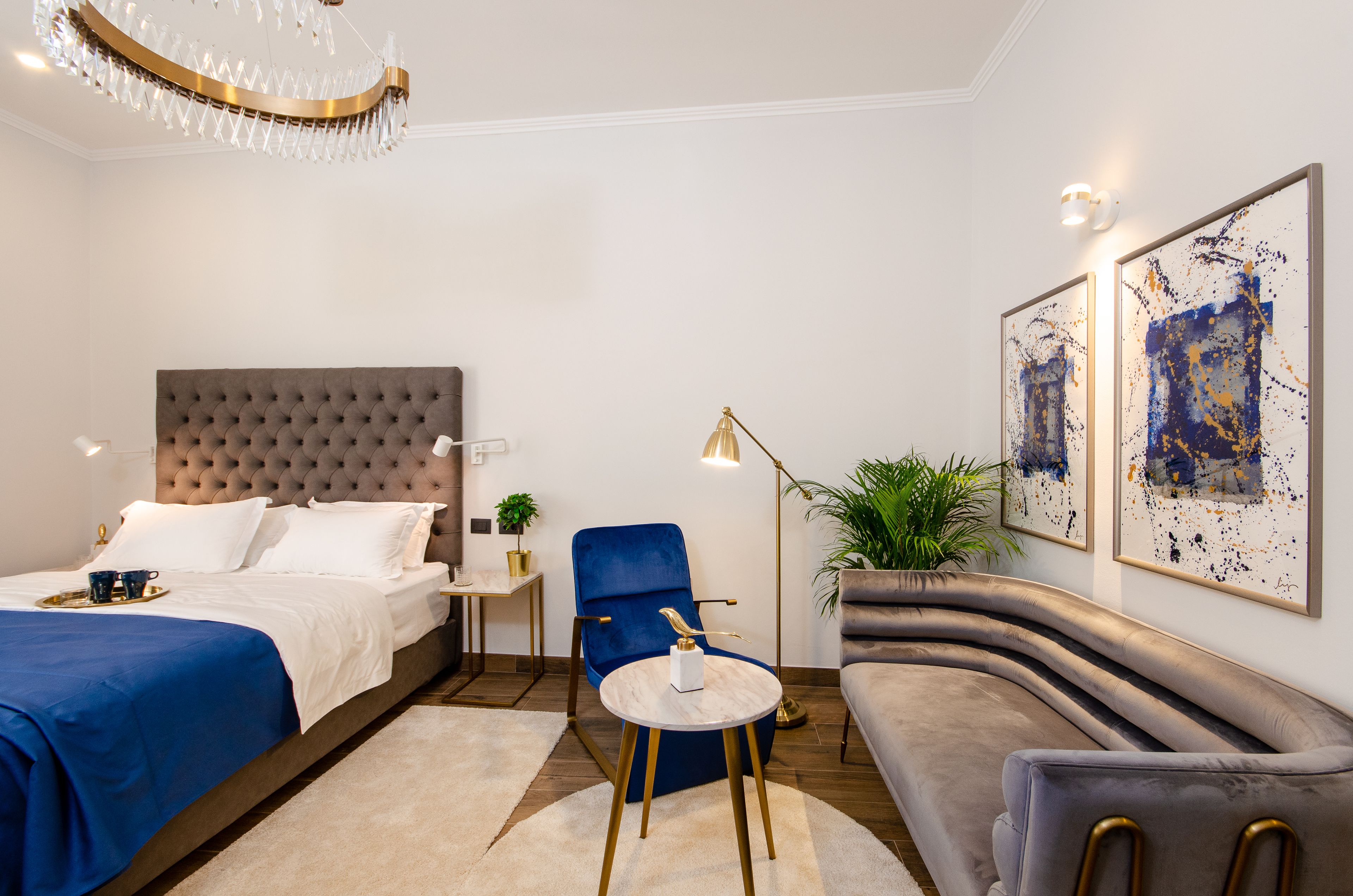 Una habitación de hotel moderna con una cama grande, un sillón azul, un sofá gris y dos cuadros abstractos en la pared.