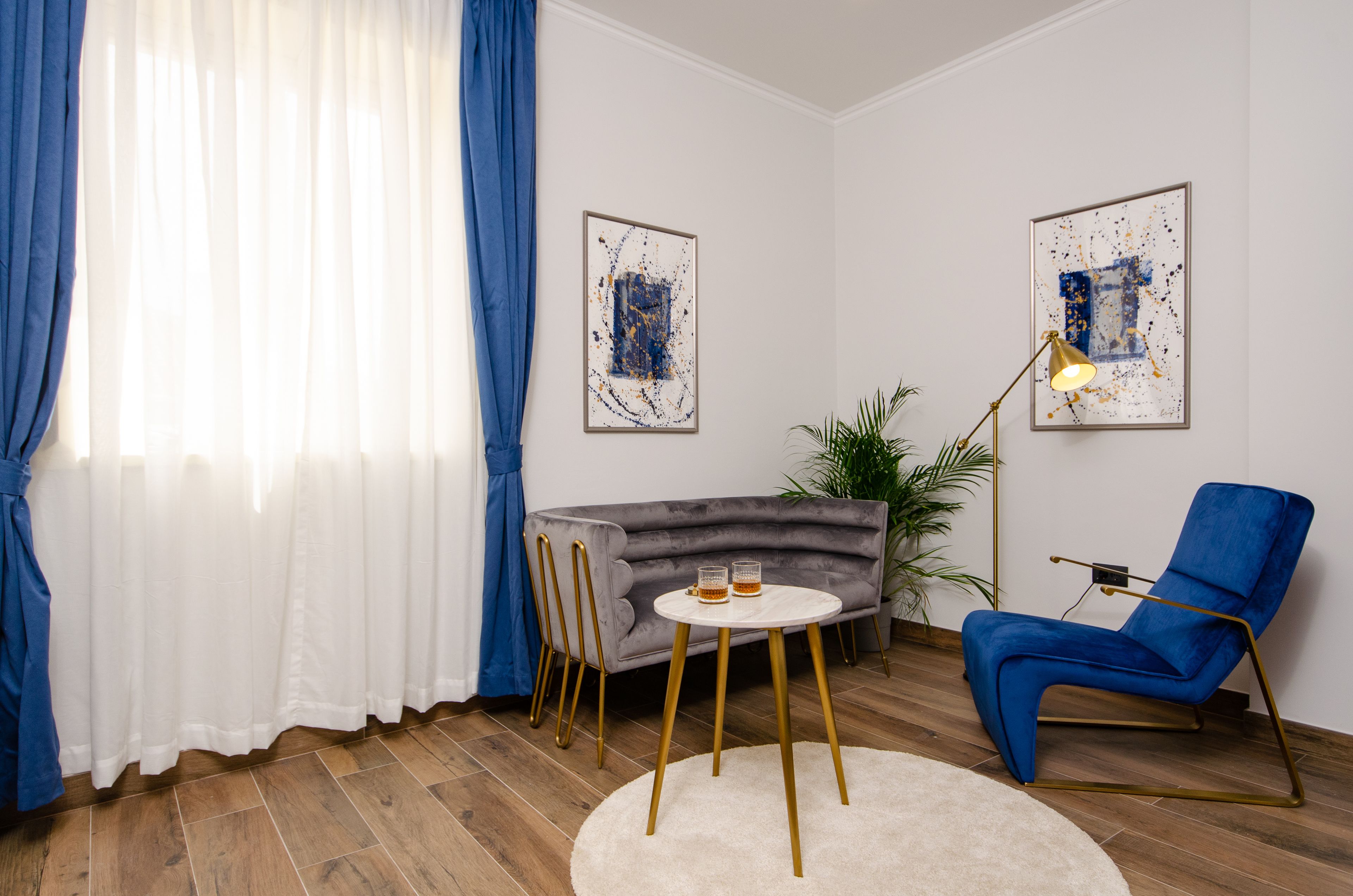 Un salon moderne avec un canapé gris, un fauteuil bleu, une table ronde avec deux verres, et deux tableaux abstraits accrochés au mur.