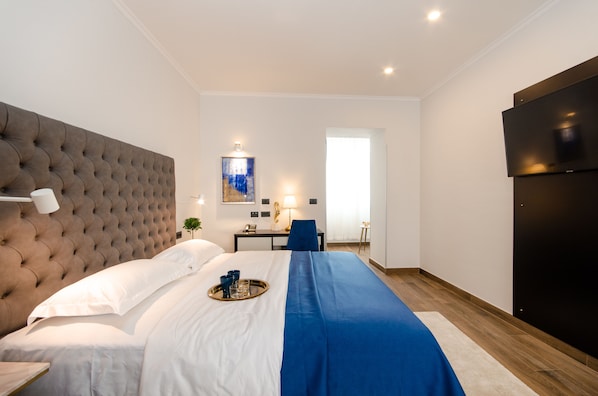 Una habitación de hotel moderna con cabecera capitonada, una cama grande con detalles en azul, un escritorio con silla, un televisor de pantalla plana y un cuadro en la pared.