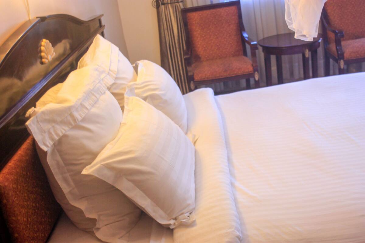 A hotel room with a bed, pillows, a wooden headboard, a small table, and two chairs.