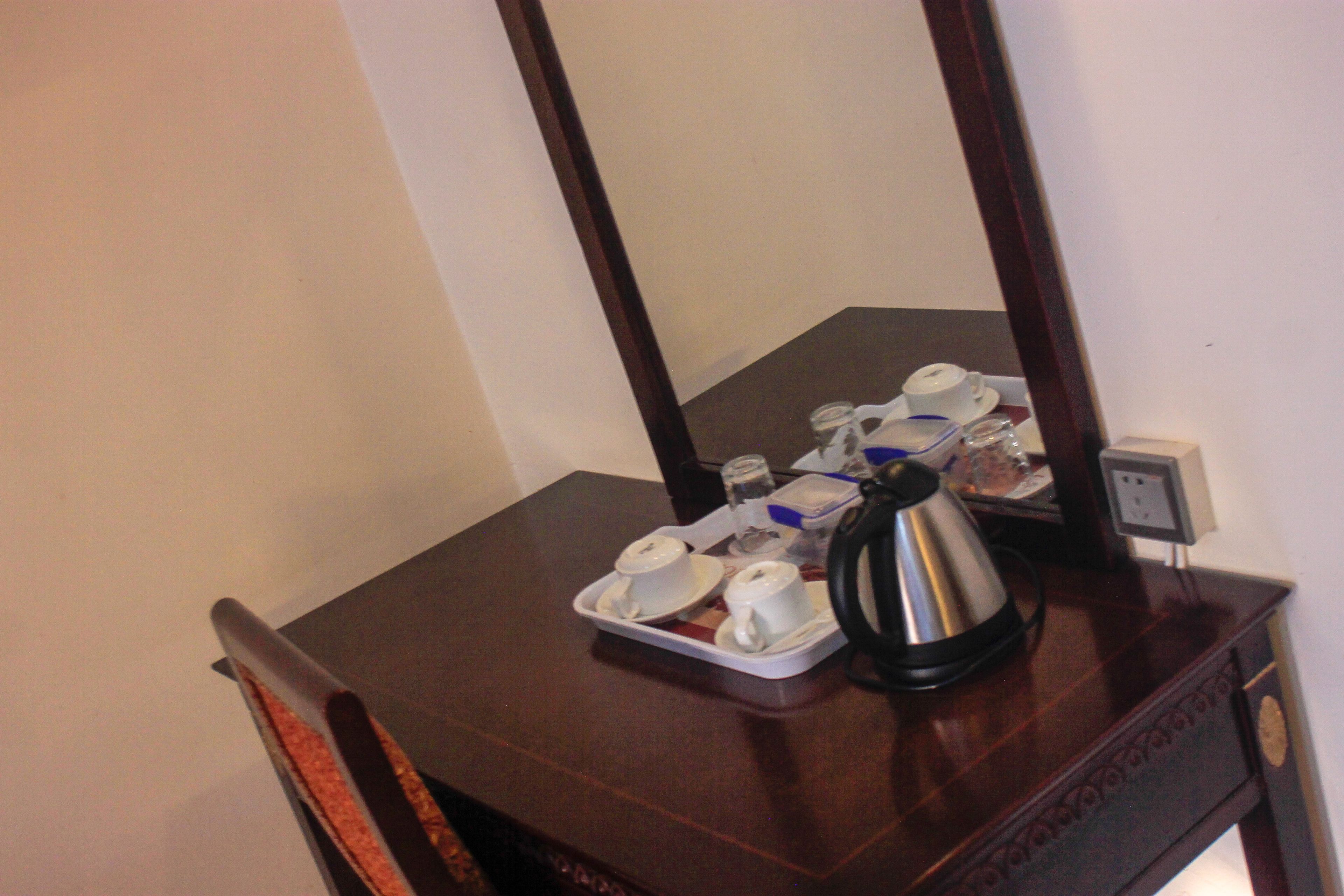 Une commode en bois sur laquelle se trouve un plateau avec des tasses et des soucoupes blanches, une bouilloire en argent et une petite prise électrique.