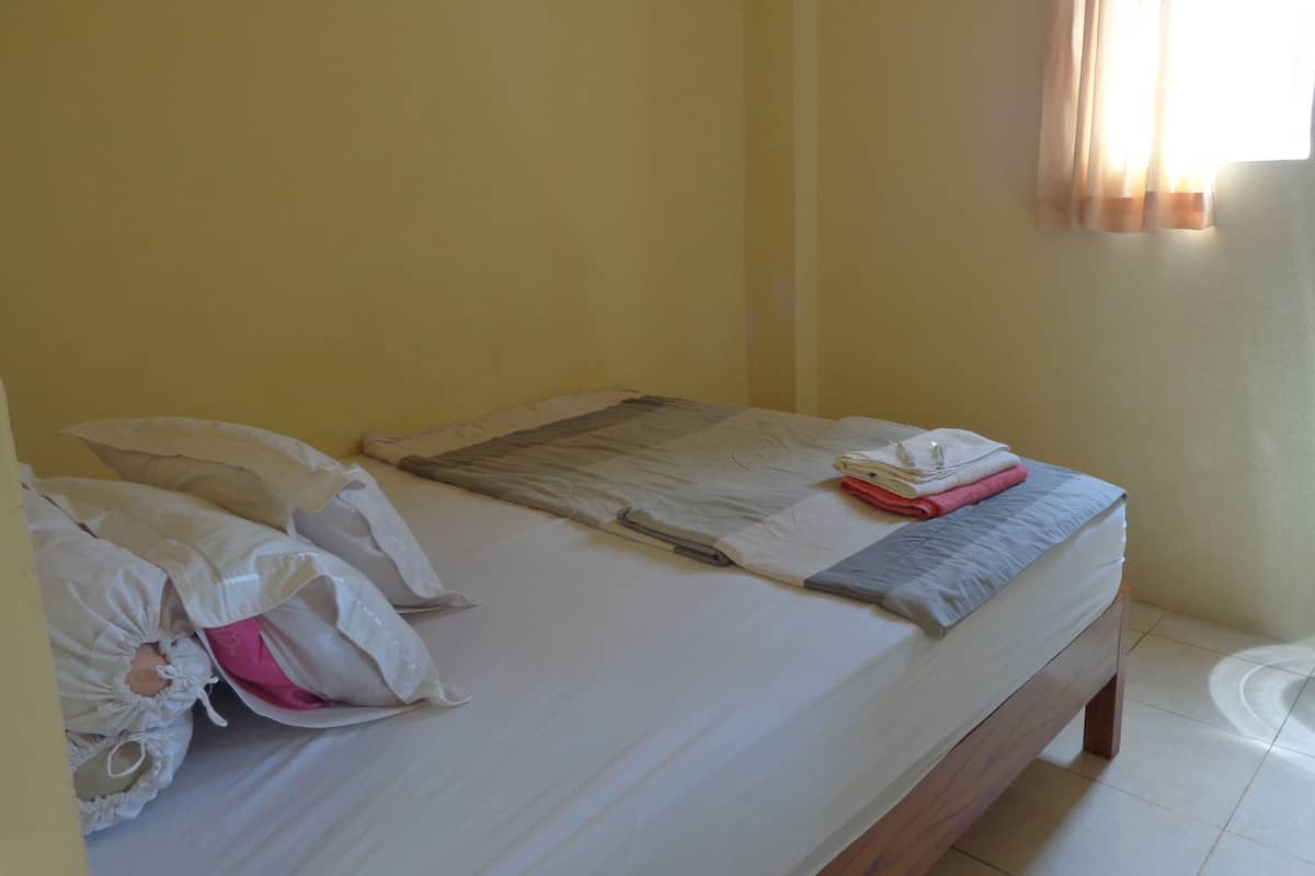 A neatly made bed with pillows and a folded towel, a wooden headboard, and a window with curtains.