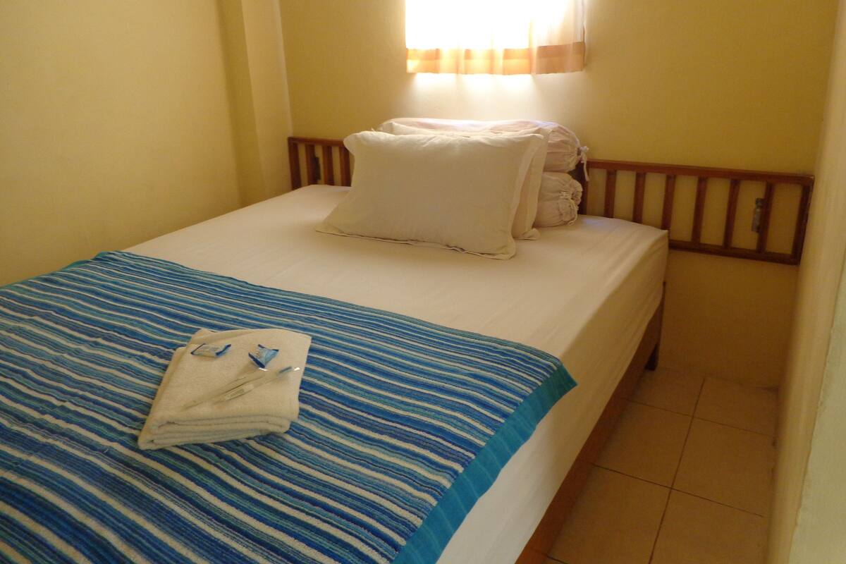 A single bed with a blue-striped bedspread and white pillows, a folded towel, and a wooden headboard.