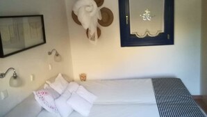 Una cama bien hecha con sábanas blancas y una manta a rayas, una mesita de noche con una lámpara, un cuadro en la pared y un adorno colgante.