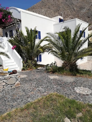 A white building with blue shutters, a stone-paved courtyard, and palm trees.