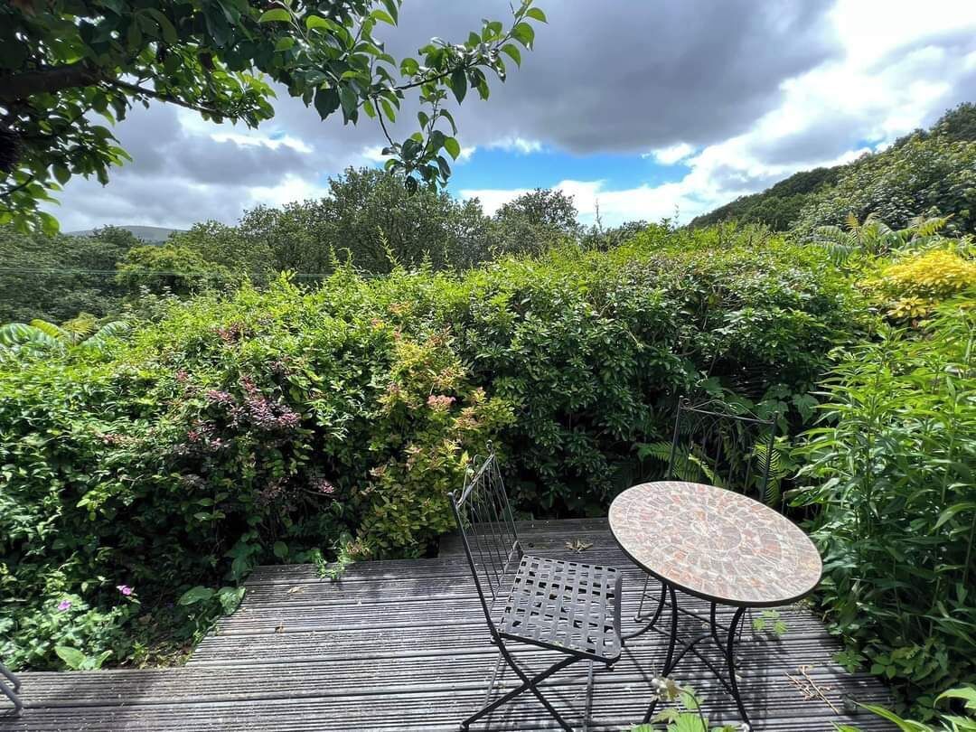 Eine Holzterrasse mit einem Metalltisch und Stuhl, umgeben von üppiger Vegetation und Hügeln im Hintergrund.