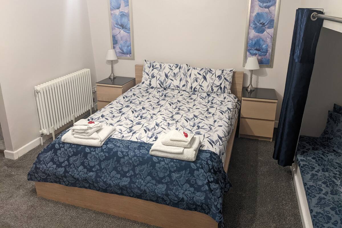 Un lit bien fait, avec une literie à motifs floraux, deux tables de chevet avec des lampes, un radiateur et deux tableaux à motifs floraux encadrés accrochés aux murs.