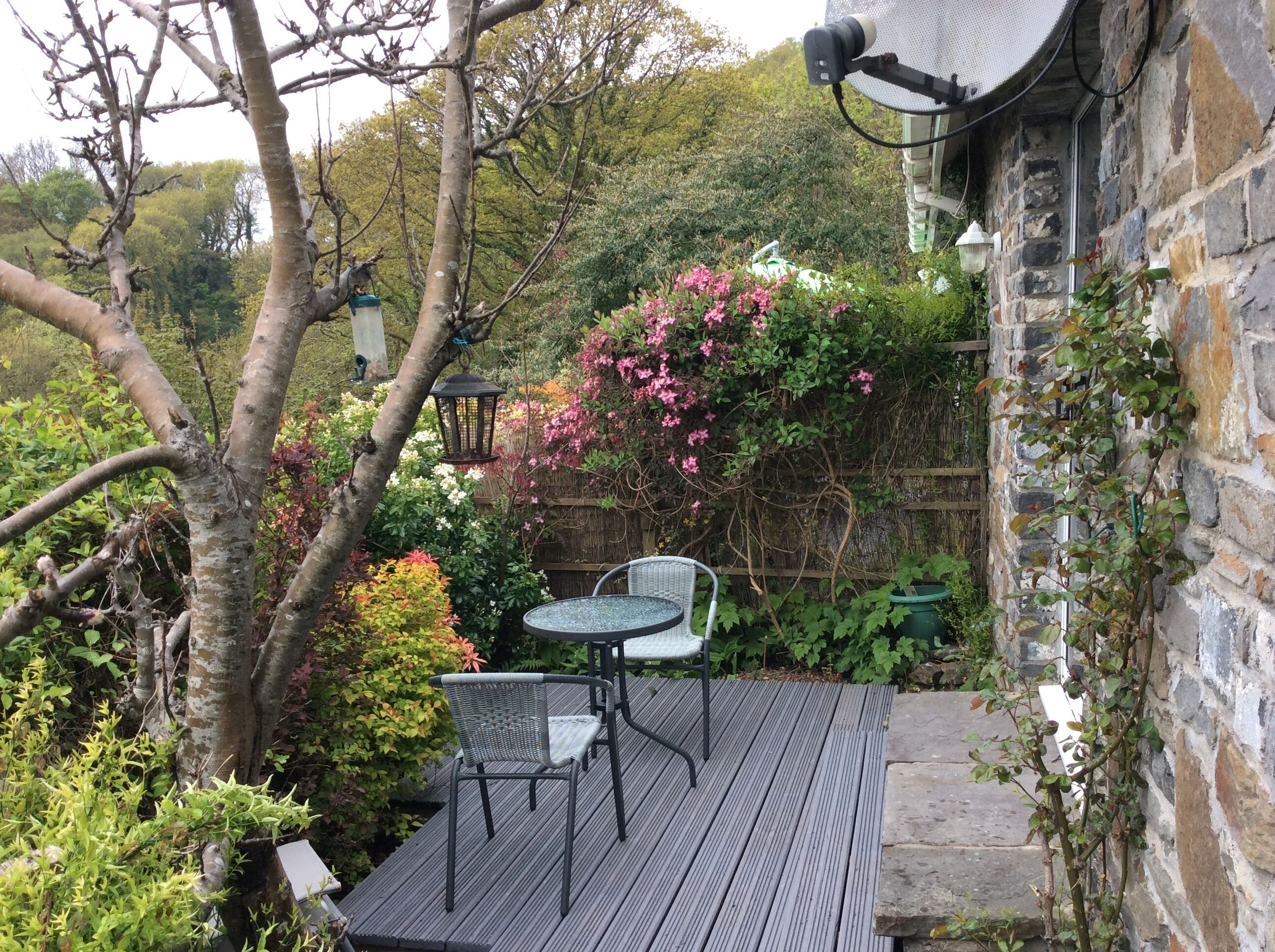 Een houten terras met twee stoelen en een tafel, omgeven door weelderige begroeiing en bloeiende planten.