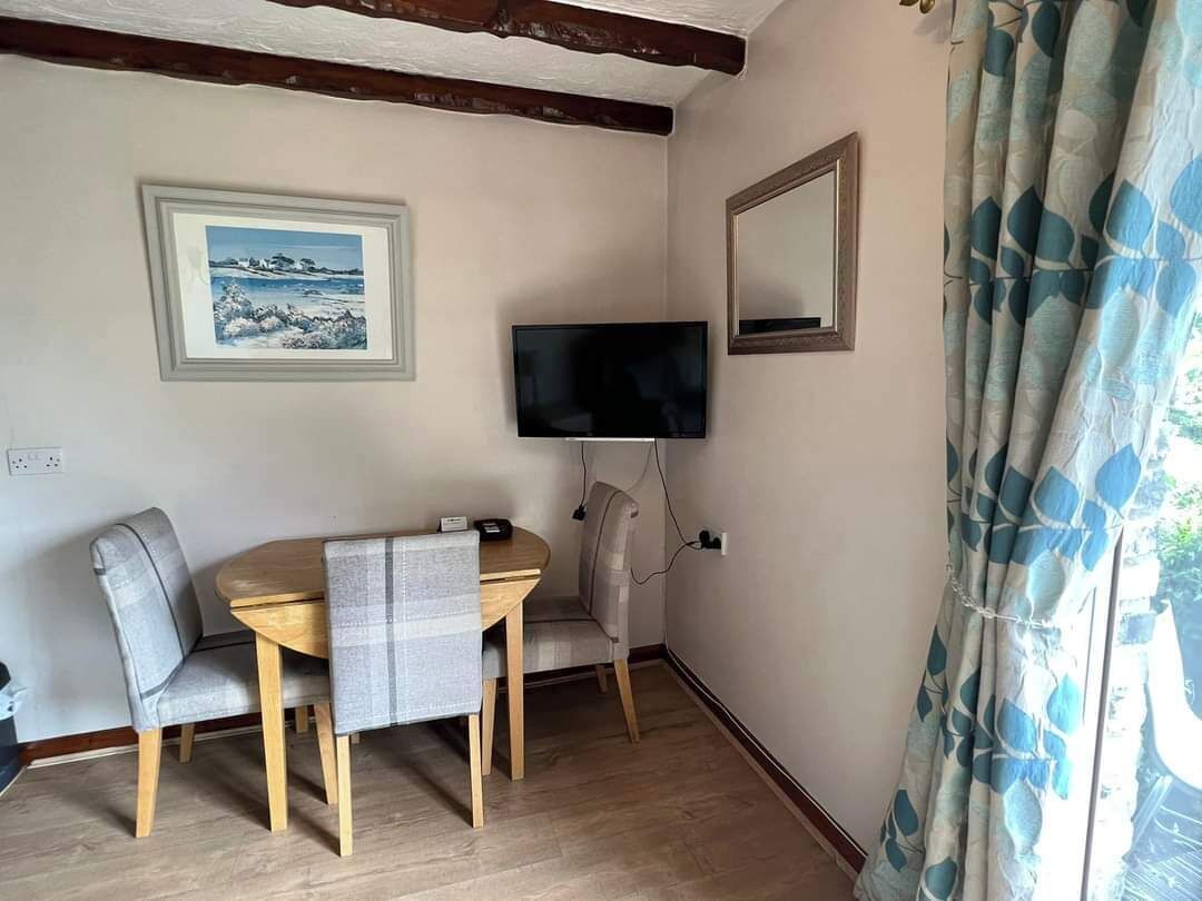 Ein Zimmer mit einem Holztisch, zwei grau gepolsterten Stühlen, einem an der Wand befestigten Fernseher und einem Fenster mit blumigem, blauem Vorhang.