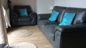A living room with a dark leather sofa and armchairs, two patterned throw pillows, a round rug, and a lamp on a side table.