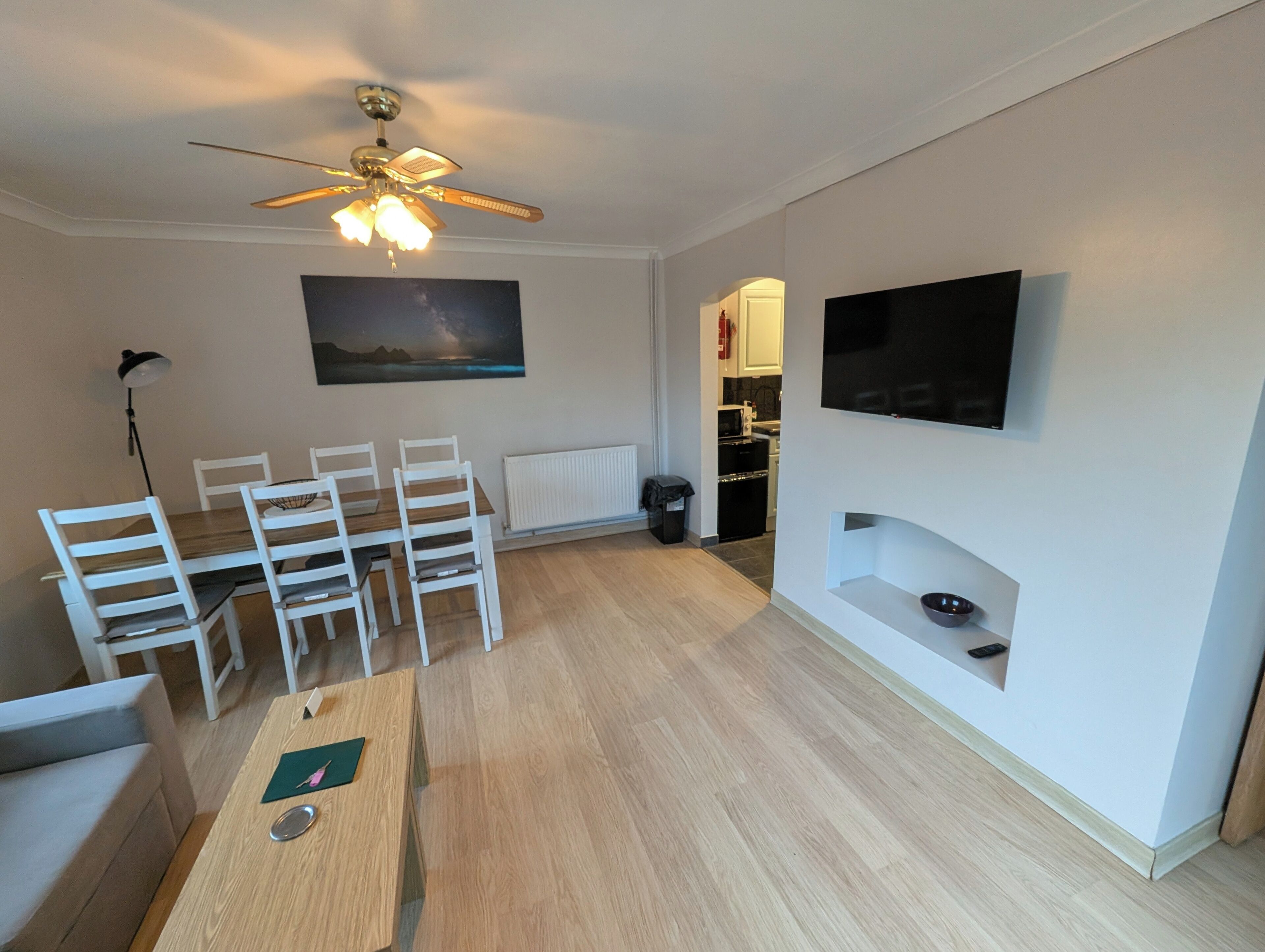 A modern living room with a dining area, a ceiling fan, a flat-screen TV mounted on the wall, and a view of the kitchen through an open doorway.