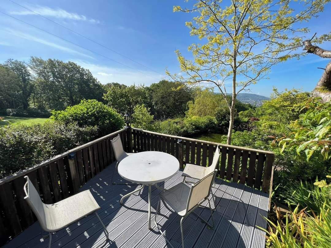 A wooden deck with a round table and two chairs, surrounded by a wooden fence, overlooking a lush garden and distant landscape.