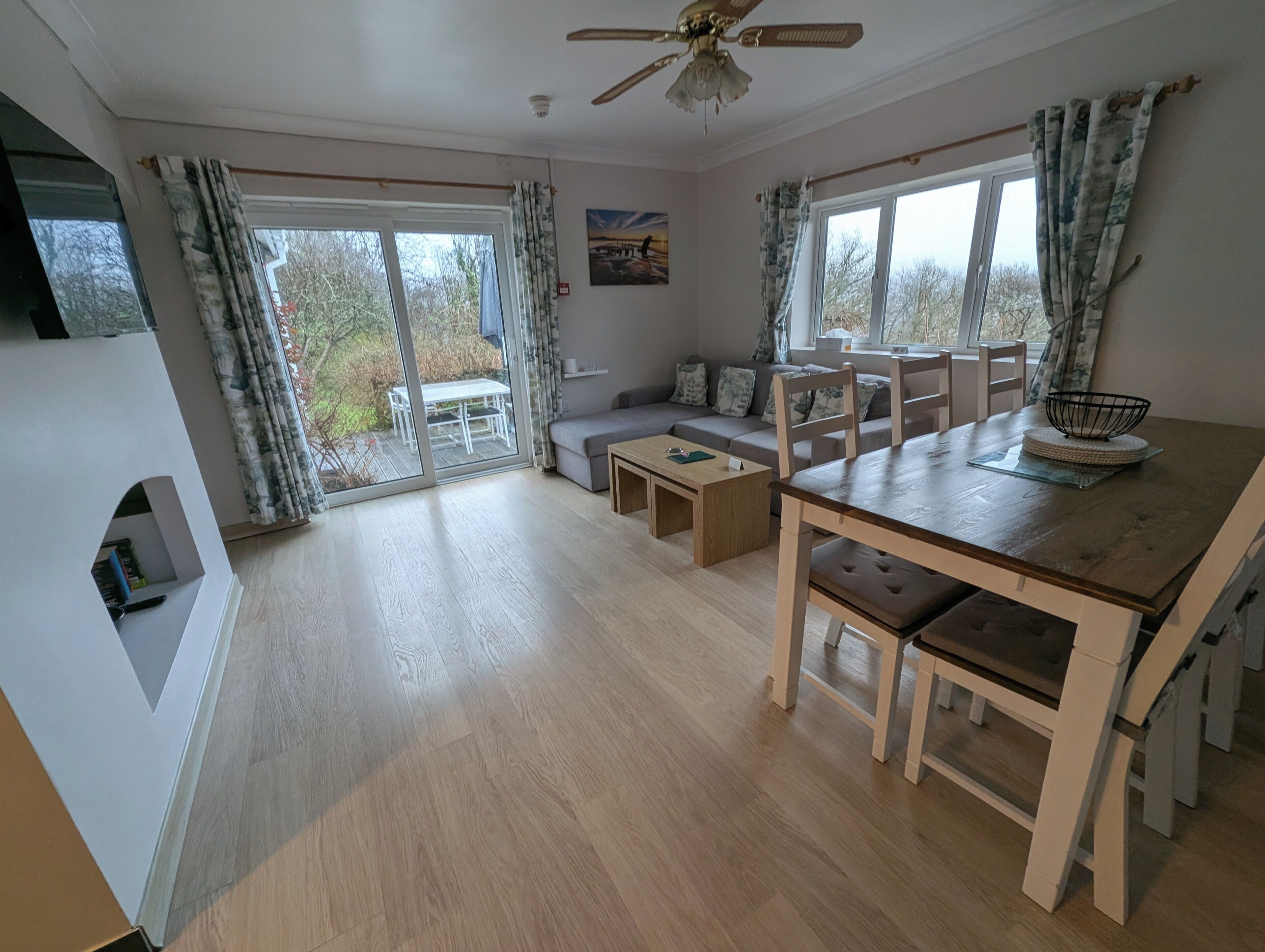 A spacious living area with a sofa, dining table, and chairs, large windows with curtains, and a ceiling fan.