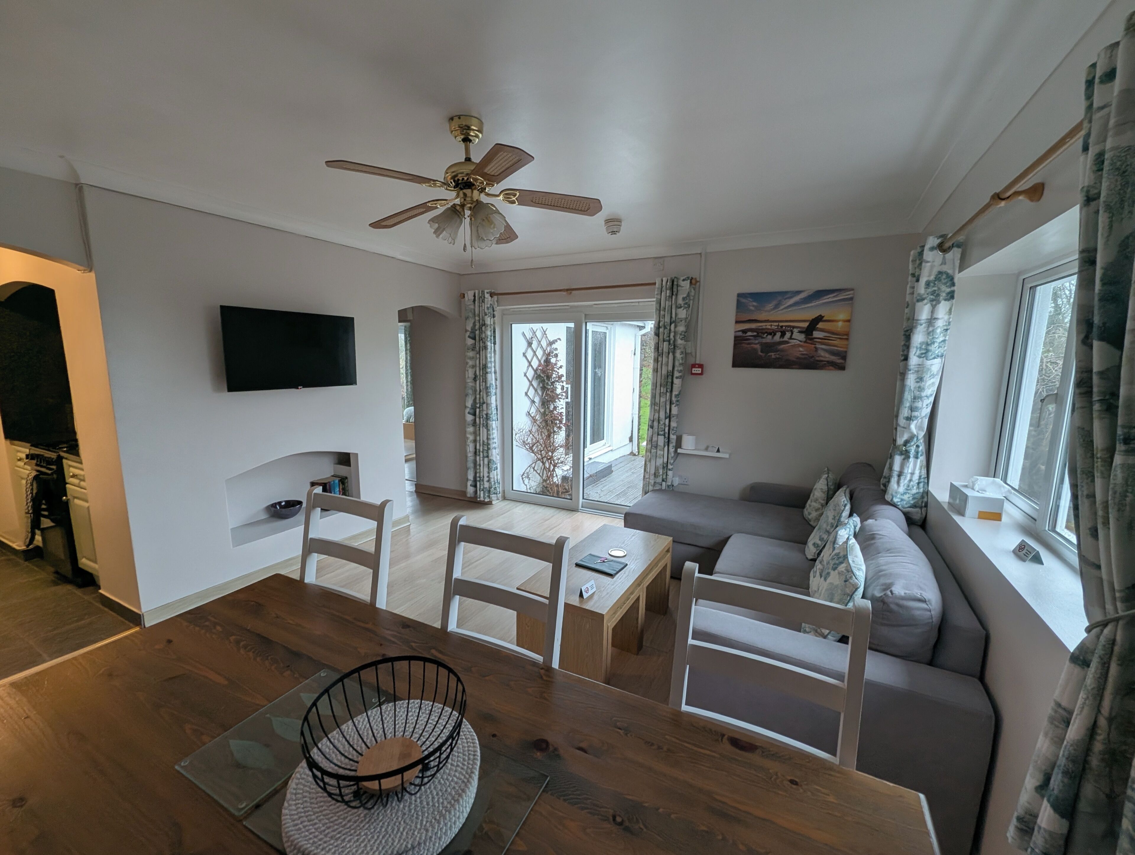 A modern living room with a ceiling fan, a flat-screen TV, a dining table with chairs, a sofa, and a window with curtains.