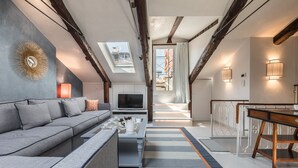 Un salon moderne avec un canapé, une table basse et un téléviseur. Il y a une lucarne et un escalier menant à un étage supérieur.