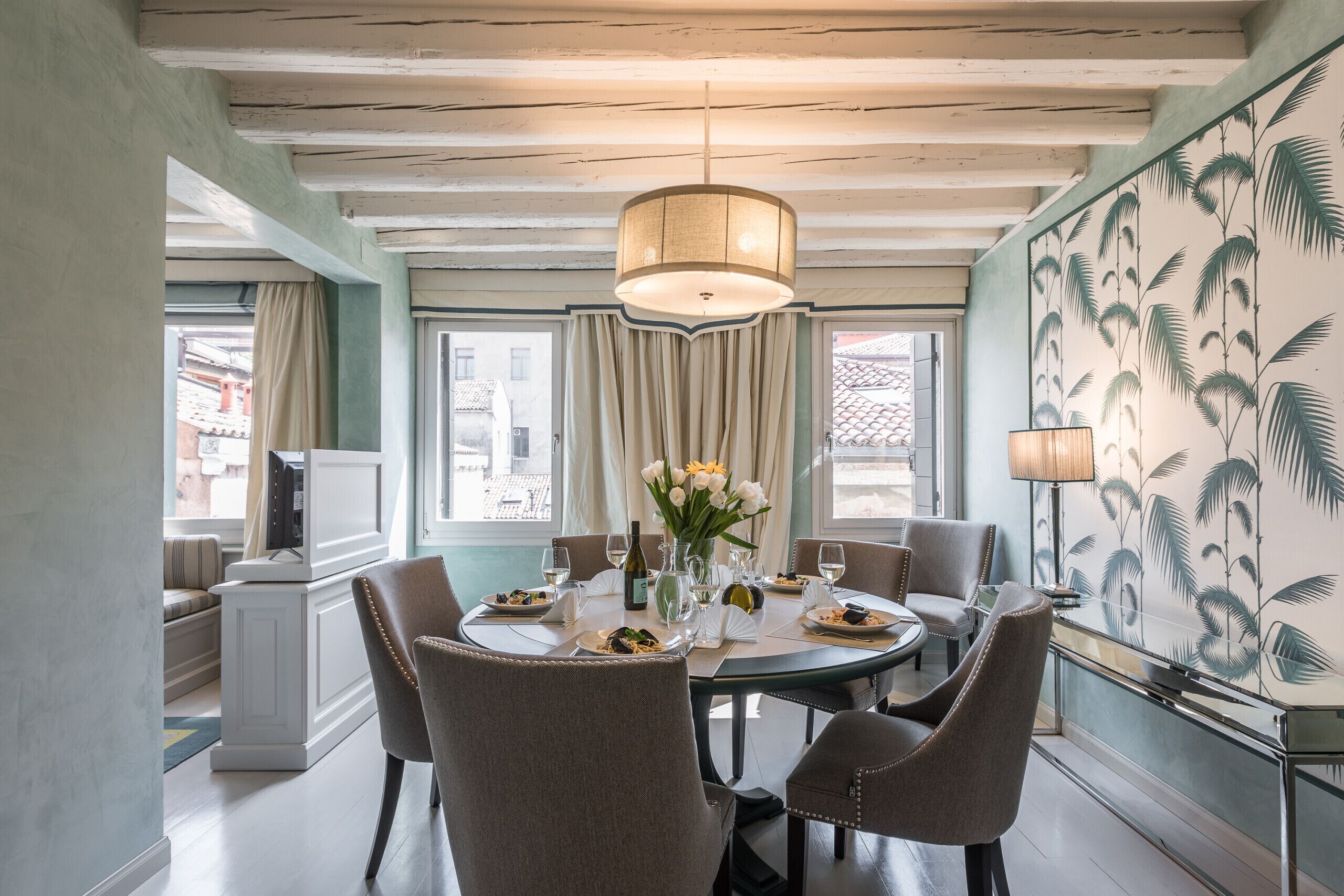 Un comedor con una mesa redonda dispuesta para cuatro personas, rodeada de sillas. Hay una araña sobre la mesa, y un jarrón con flores en ella. La habitación tiene un ventanal con cortinas, y una pared con un estampado de hojas tropicales.