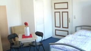 A small, simple room with a bed, a table with a chair, and a door. - Gästehaus Harz (Goslar)