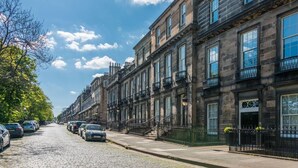 A row of historic stone townhouses with uniform architecture, a cobblestone street, and parked cars.