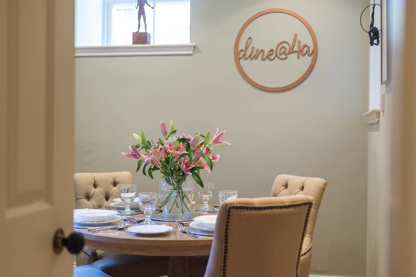 Un comedor con una mesa redonda preparada para cuatro personas, con un jarrón de lirios rosados y un letrero en la pared que dice "dine@la".