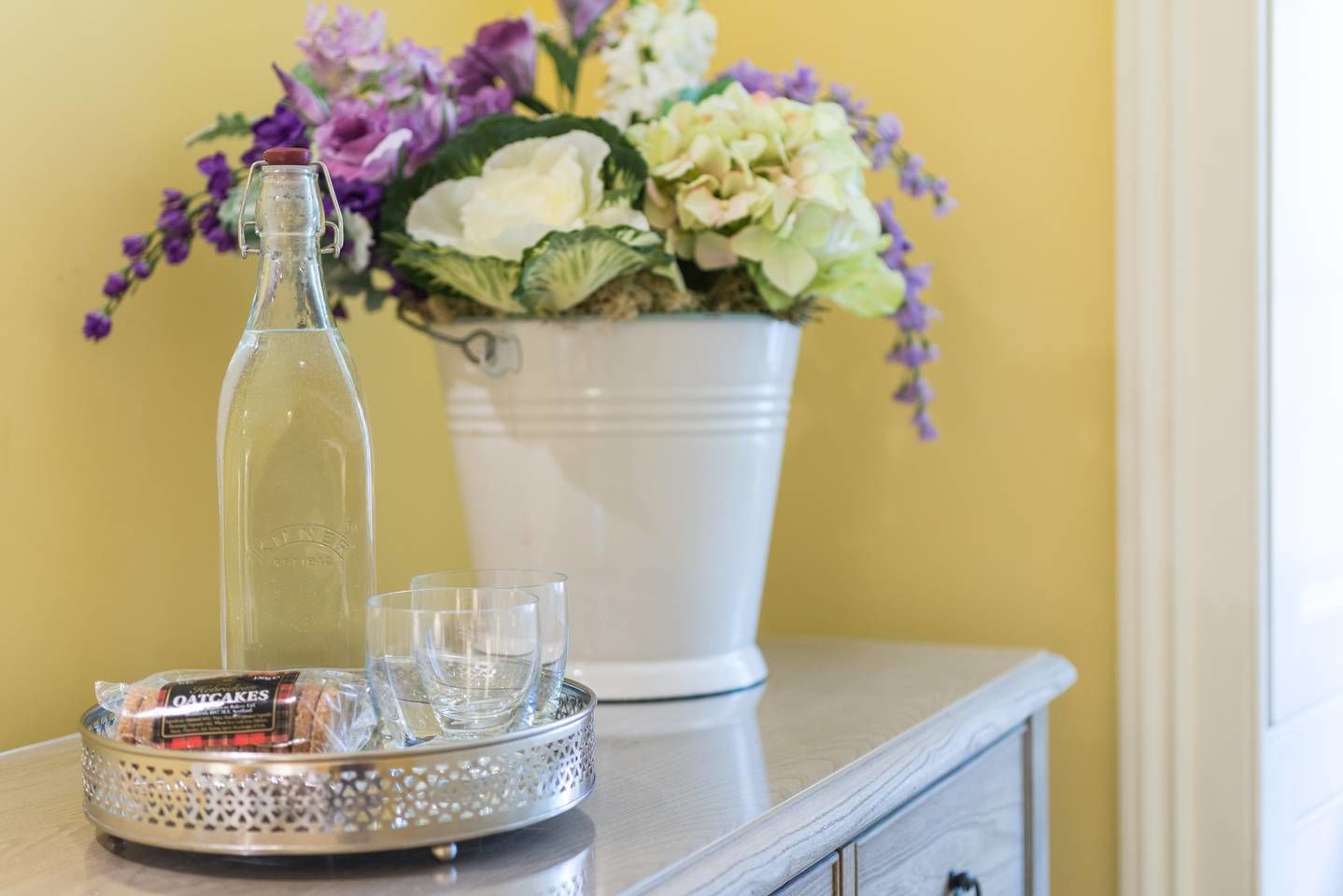 Eine klare Glasflasche, ein weißer Eimer mit Blumen und ein Tablett mit einer verpackten Ware und Gläsern auf einer Holzoberfläche.