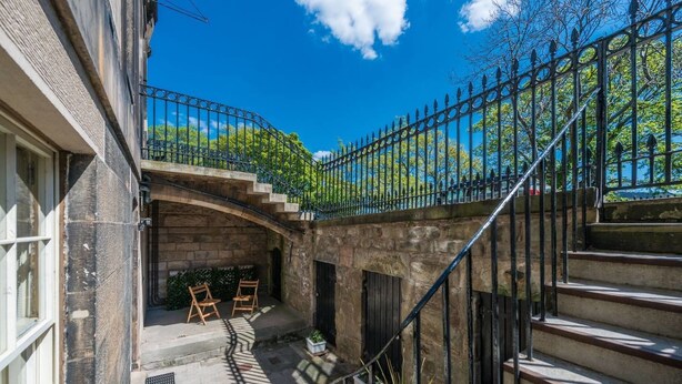 A stone staircase leading to a balcony with a wrought iron railing, a small seating area with chairs, and a view of trees and sky.