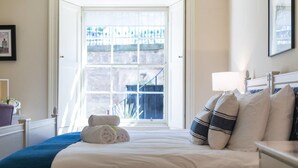 A bedroom with a large window, a bed with white linens, a bedside table with a lamp, and a framed picture on the wall.