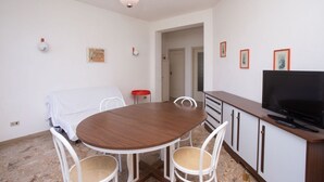 Een eetruimte met een ronde tafel, witte stoelen, een televisie en een kast.