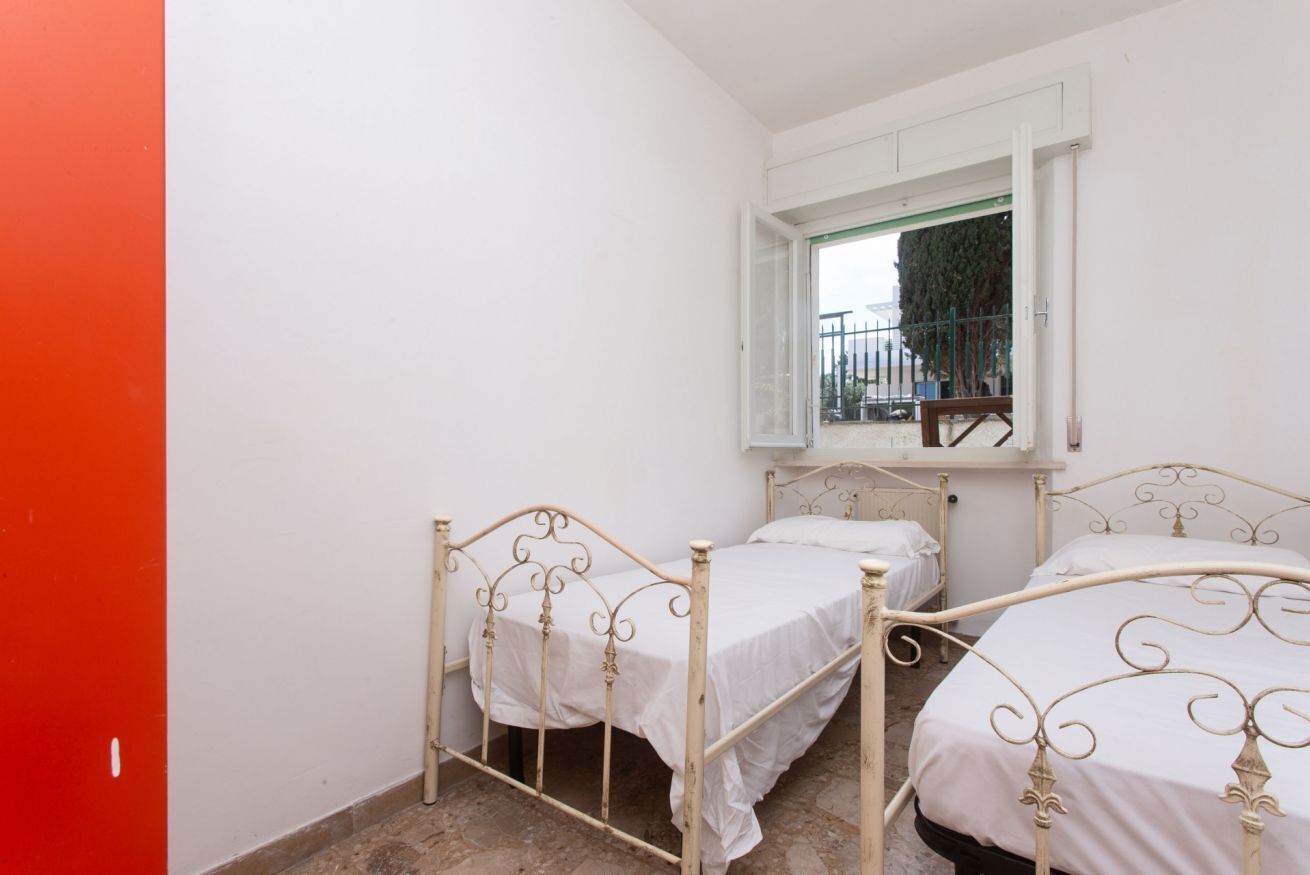 A small, white-walled room with two metal beds and a window overlooking a balcony.