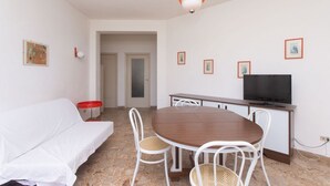 Een kamer met een witte bank, een houten eettafel met stoelen, een televisie en lijstjes met foto's aan de muur.