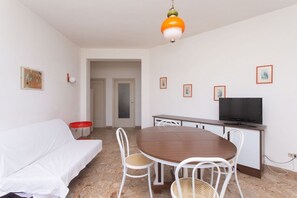 Ein Zimmer mit einer weißen Couch, einem Holzesstisch mit Stühlen, einem Fernseher und gerahmten Bildern an der Wand.