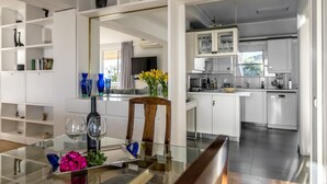 A modern kitchen with white cabinets, a dining area with a glass table, and a view into the living room. - Amazing Beach Front Apartment in the Athens Riviera (Vouliagmeni)