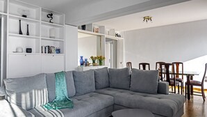 A modern living room with a grey sectional sofa, white shelving units, and a dining area with wooden chairs and a glass table. - Amazing Beach Front Apartment in the Athens Riviera (Vouliagmeni)
