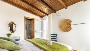 A bedroom with a wooden ceiling, a bed with a green blanket, a hat on the bed, a mirror, a hat on the wall, and a chair. - Villa Sun Total Relax (Alcamo)
