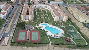 Luftaufnahme eines Resorts mit einem zentralen Schwimmbad, Tennisplätzen und einer angelegten Fläche mit Palmen.