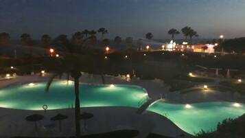 Vista nocturna de un resort con dos piscinas iluminadas rodeadas de palmeras y un edificio distante con luces.