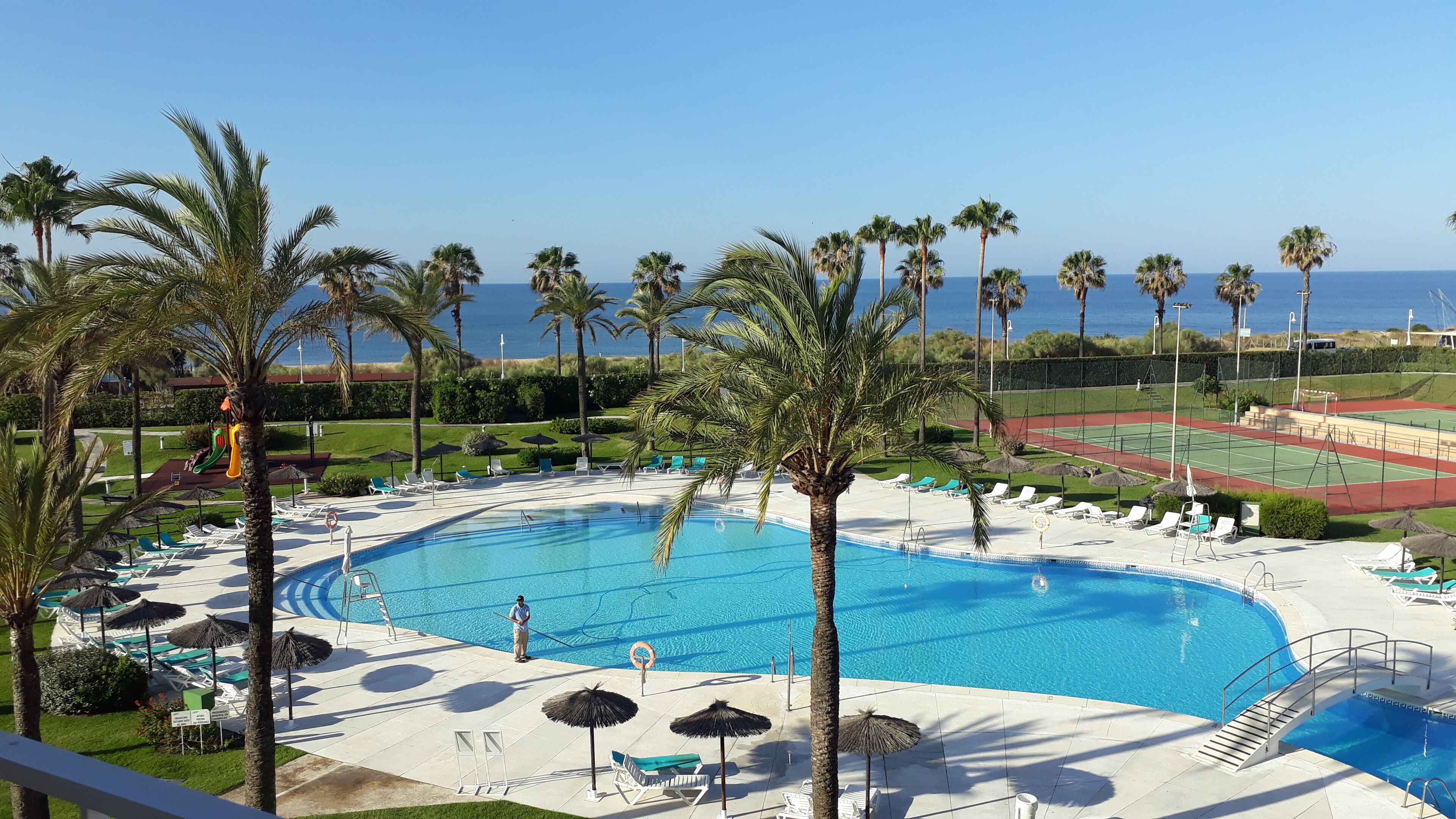 Piscina con tumbonas y palmeras, rodeada de una cancha de tenis y vista al mar.