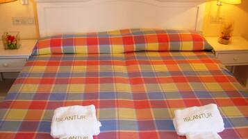Een bed met een kleurrijk geruit sprei, twee handdoeken met de tekst 'ISLANDUR' en bedlampjes.