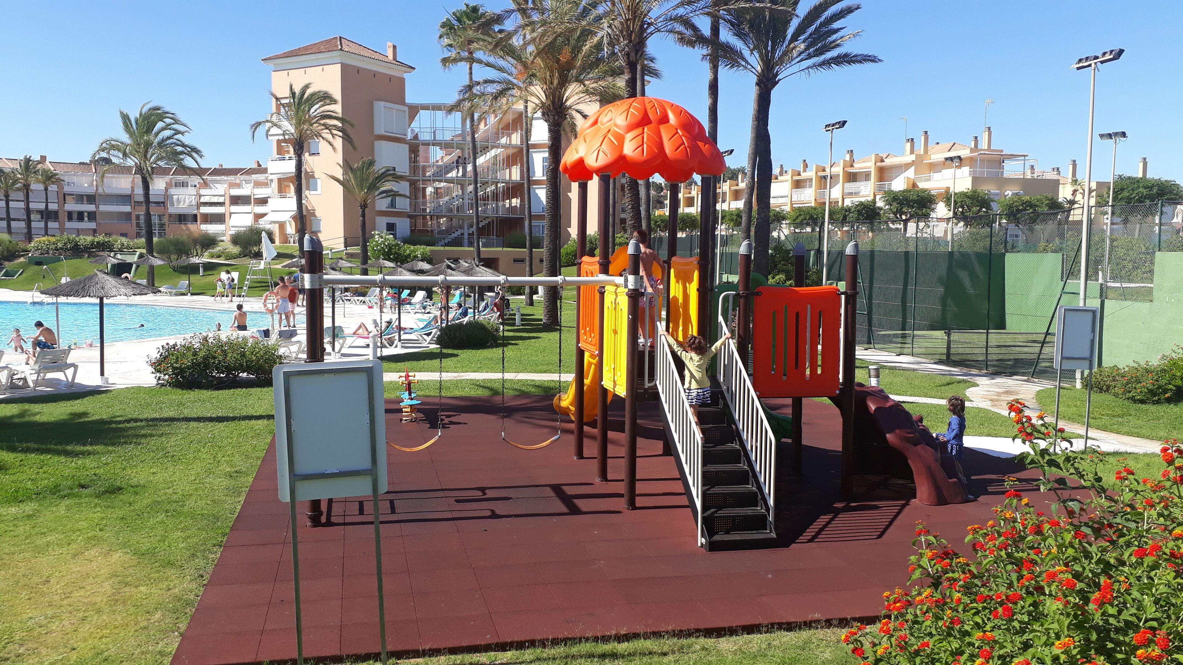 A playground with swings and slides, a pool area with lounge chairs, and a building complex in the background.