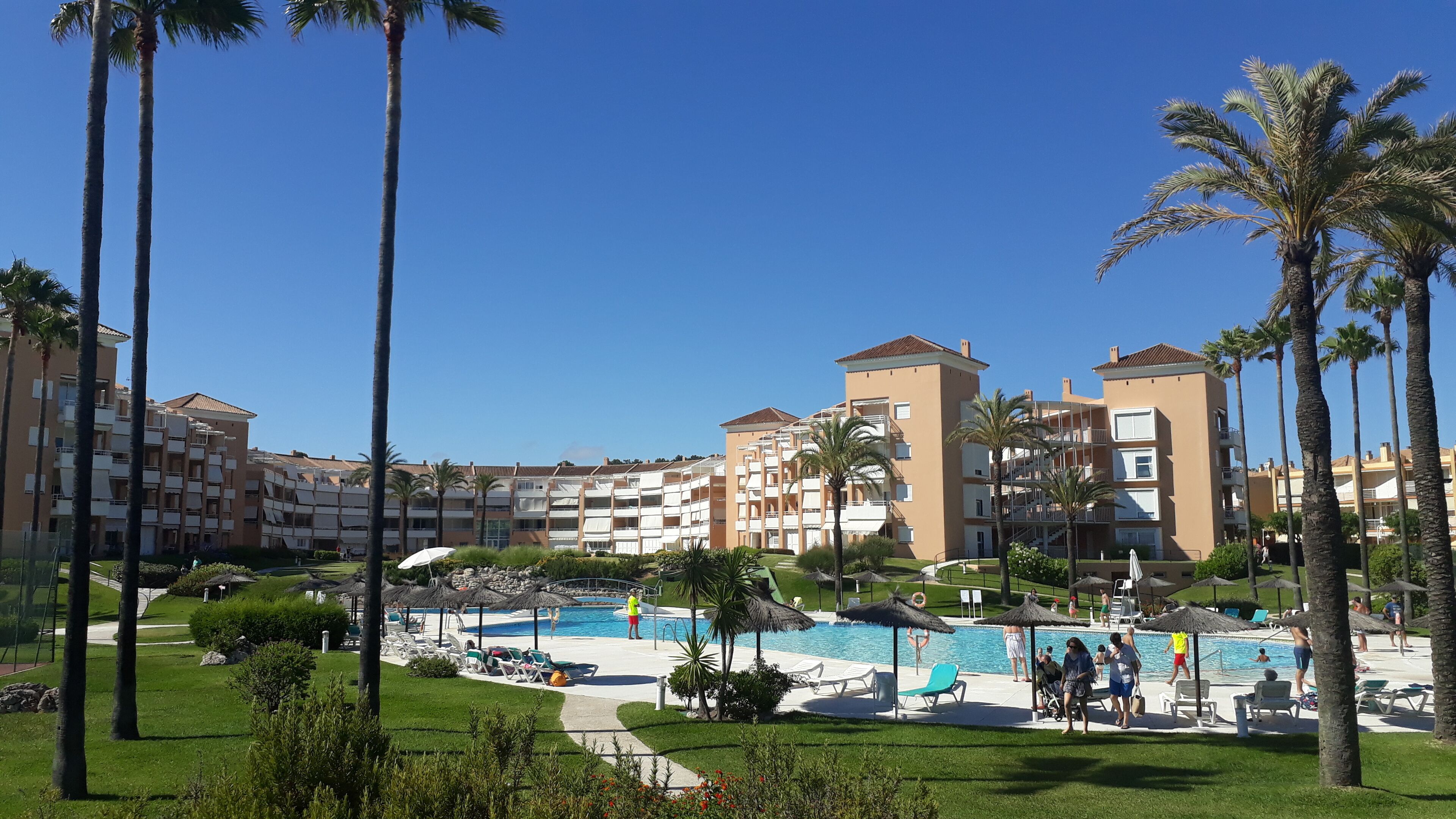 Ein Resort mit mehrstöckigen Gebäuden, einem Schwimmbeckenbereich inklusive Liegestühlen und Sonnenschirmen sowie Palmen.