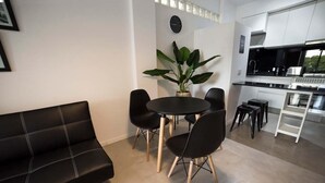 A compact living space with a black sofa, a round dining table with four chairs, a potted plant, and a kitchenette with modern appliances.