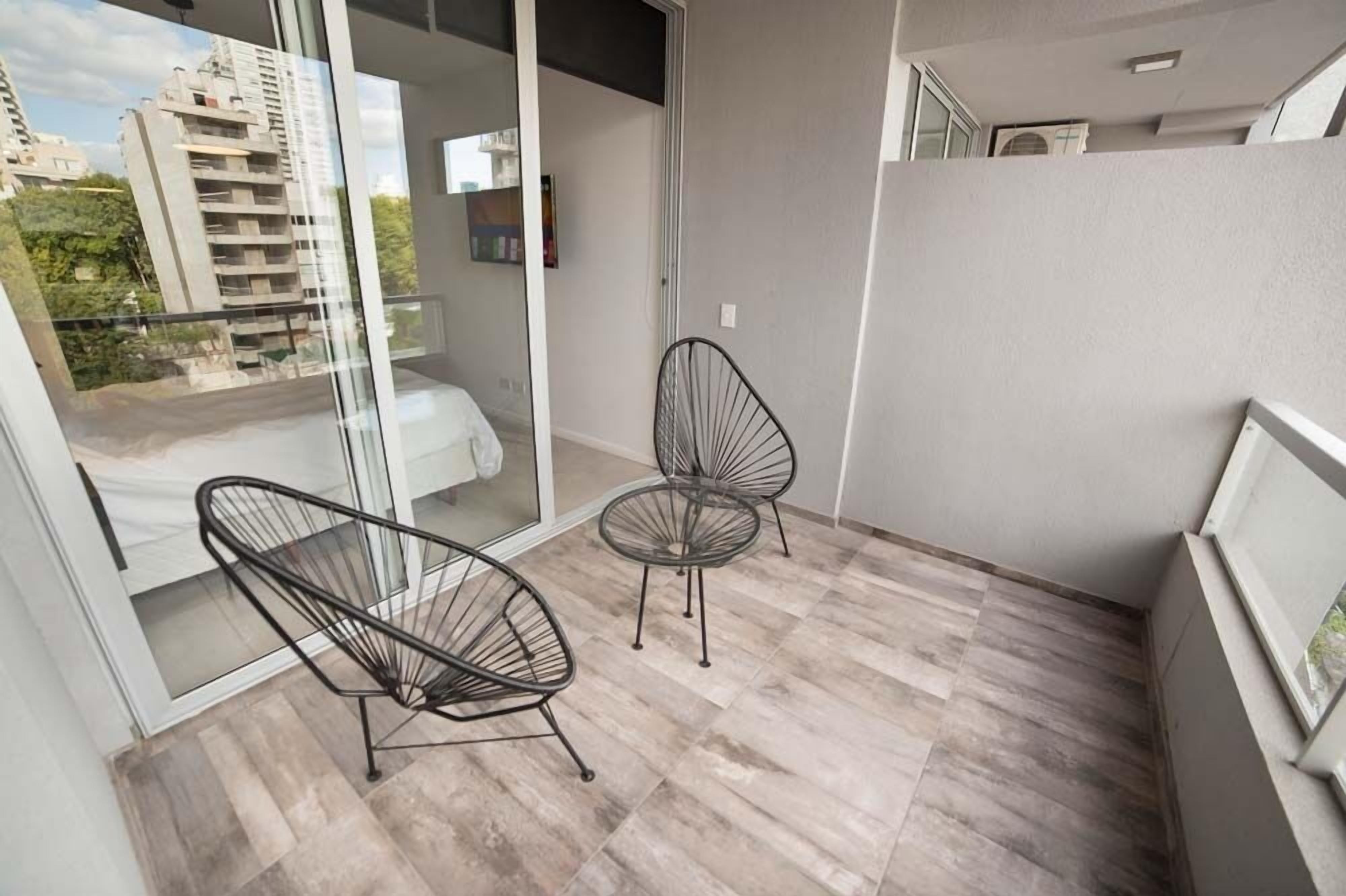 A balcony with two black wireframe chairs, a glass door leading to a bedroom, and a view of a high-rise building.