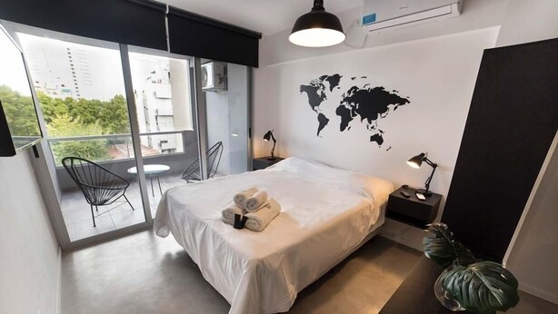 A modern hotel room with a large bed, a world map on the wall, a balcony with a chair, and a potted plant.
